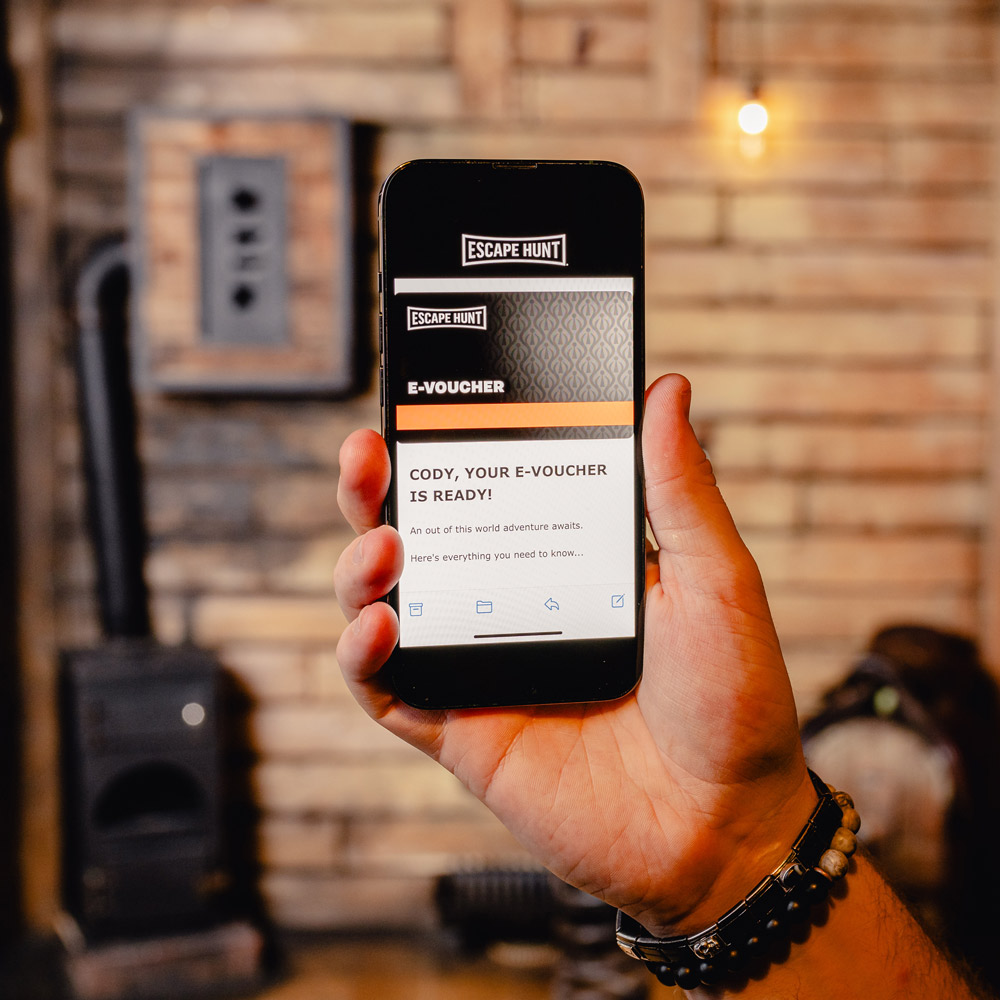 A man's hand holding a mobile phone showing the Escape Hunt e-voucher page